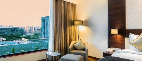A hotel room with a large window, a bed, a chair, a small table, and a nightstand with a lamp.
