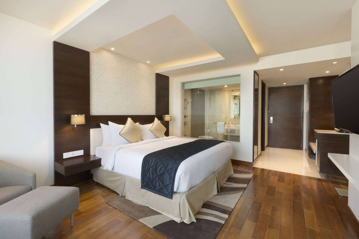 Une chambre d’hôtel moderne dotée d’un grand lit, de tables de chevet, d’un coin salon et d’une salle de bain visible à travers une porte en verre.