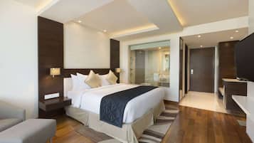 Una habitación de hotel moderna con una cama grande, mesitas de noche, una zona de estar y un baño visible a través de una puerta de vidrio.