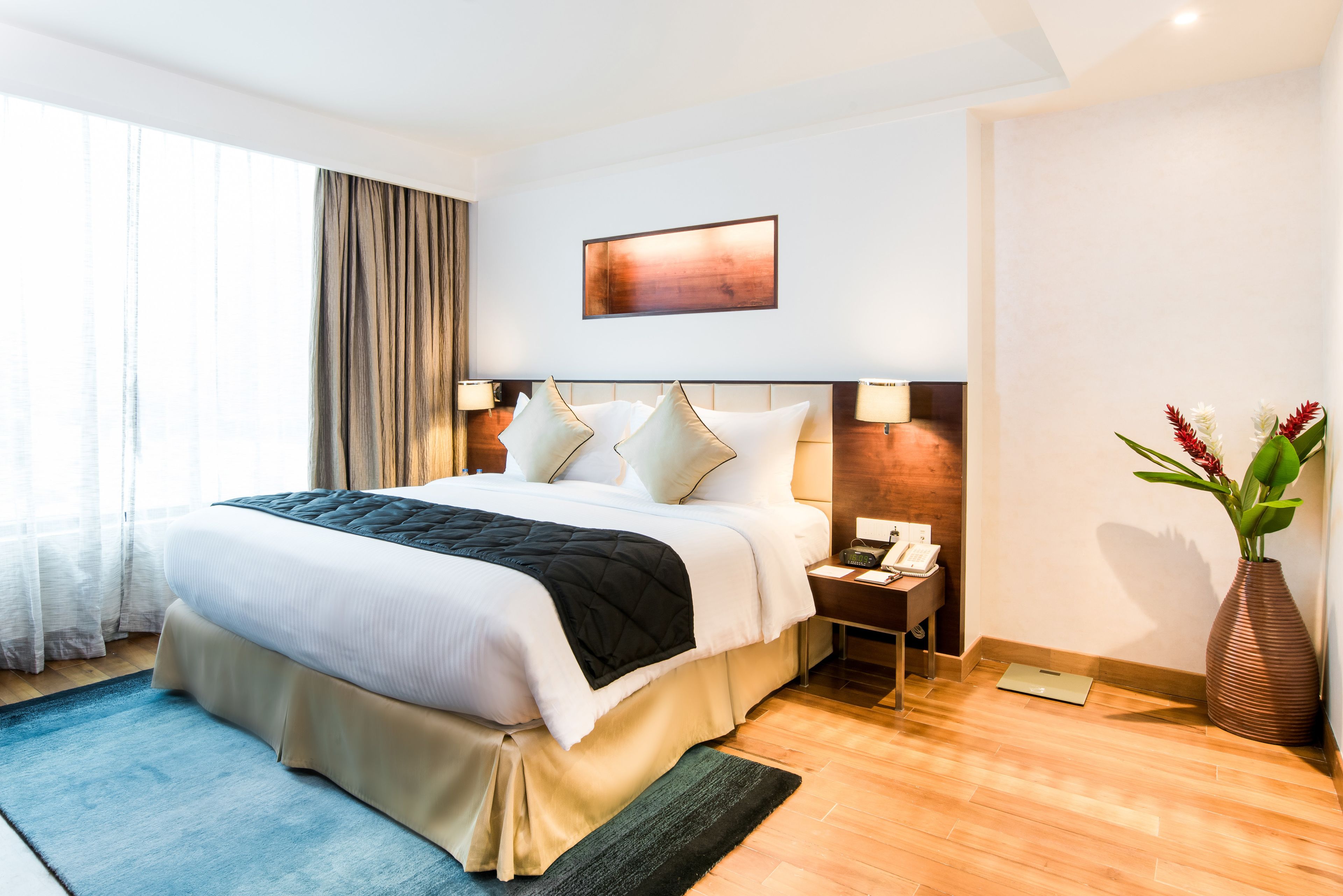 Une chambre d’hôtel avec un grand lit, des tables de chevet, une petite table, un vase avec des fleurs et un tableau au mur.