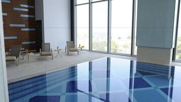 An indoor swimming pool with clear blue tiles, a wooden wall with blue accents, and a seating area with chairs and a small table.