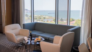 Et hotelværelse med et stort vindue, der giver udsigt over havet og en by i det fjerne. Der er en grå sofa, to lænestole og et lille bord med en flaske, glas og en skål med frugt.
