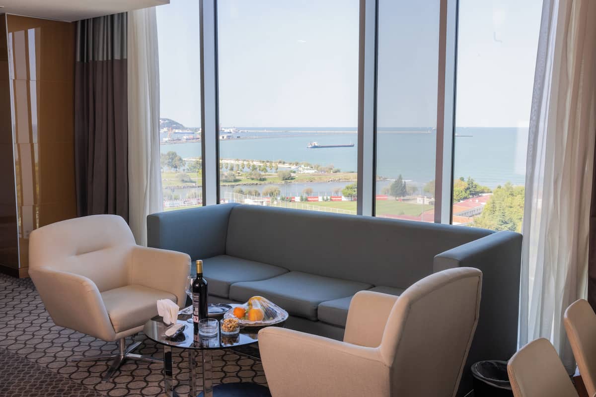 Une chambre d’hôtel dotée d’une grande fenêtre donnant sur la mer et sur le paysage urbain au loin. On y trouve un canapé gris, deux fauteuils et une petite table sur laquelle sont posés une bouteille, des verres et un panier de fruits.