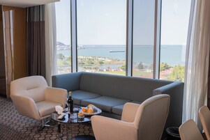 Quarto de hotel com uma grande janela que oferece vista para o mar e um horizonte urbano distante. Há um sofá cinzento, duas poltronas e uma pequena mesa com uma garrafa, copos e uma fruteira.