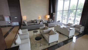 A spacious lobby with multiple seating areas, including sofas and armchairs, arranged around a central coffee table. There are two lamps on a side table, and a large window provides a view of the outside.