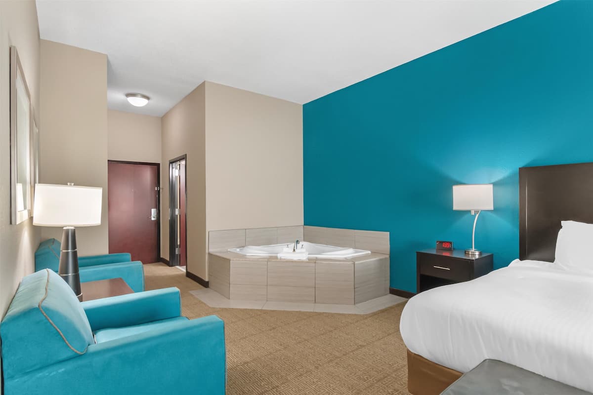 A hotel room with a large bed, a sitting area with a blue sofa, a small table, a wall-mounted TV, and a bathroom with a bathtub and a shower.