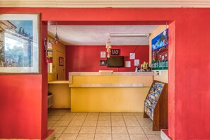Een receptiegedeelte met een rode muur, een gele balie, een brandblusser, een televisietoestel en een tijdschriftrek.
