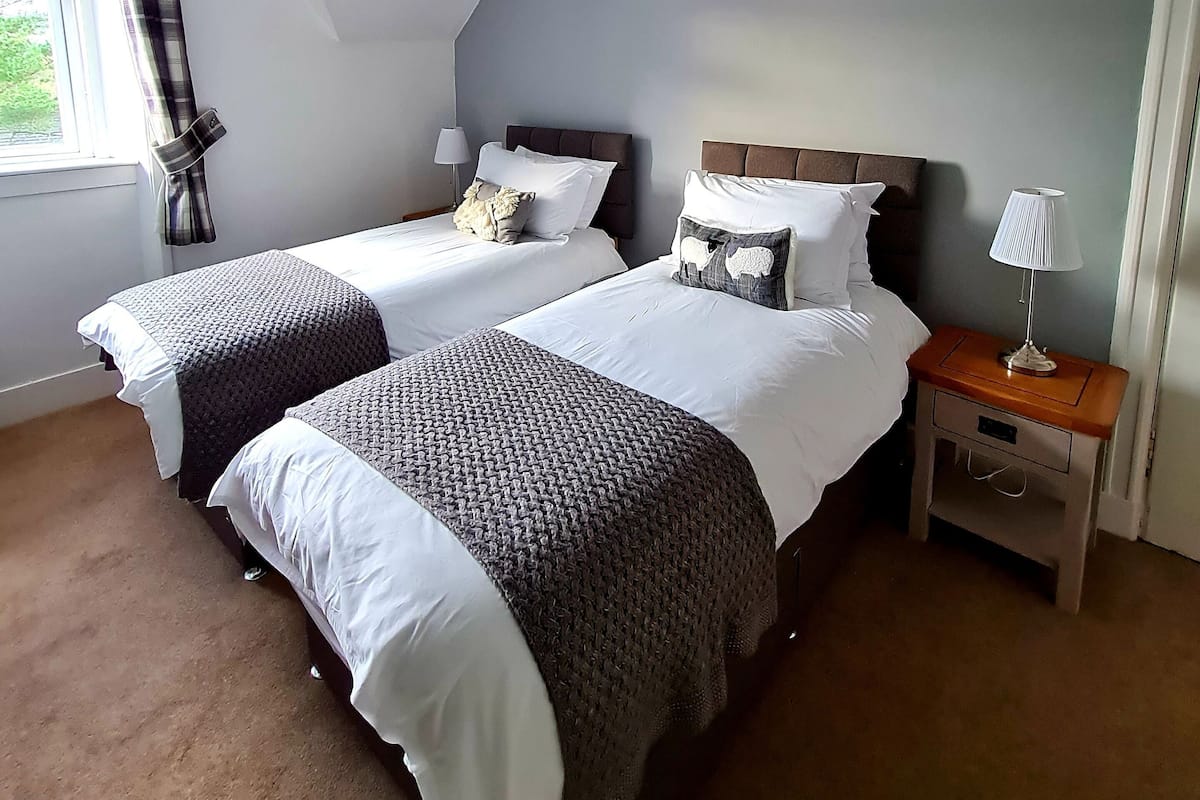 A bedroom with two beds, a nightstand, a lamp, a window with curtains, and a sloped ceiling.