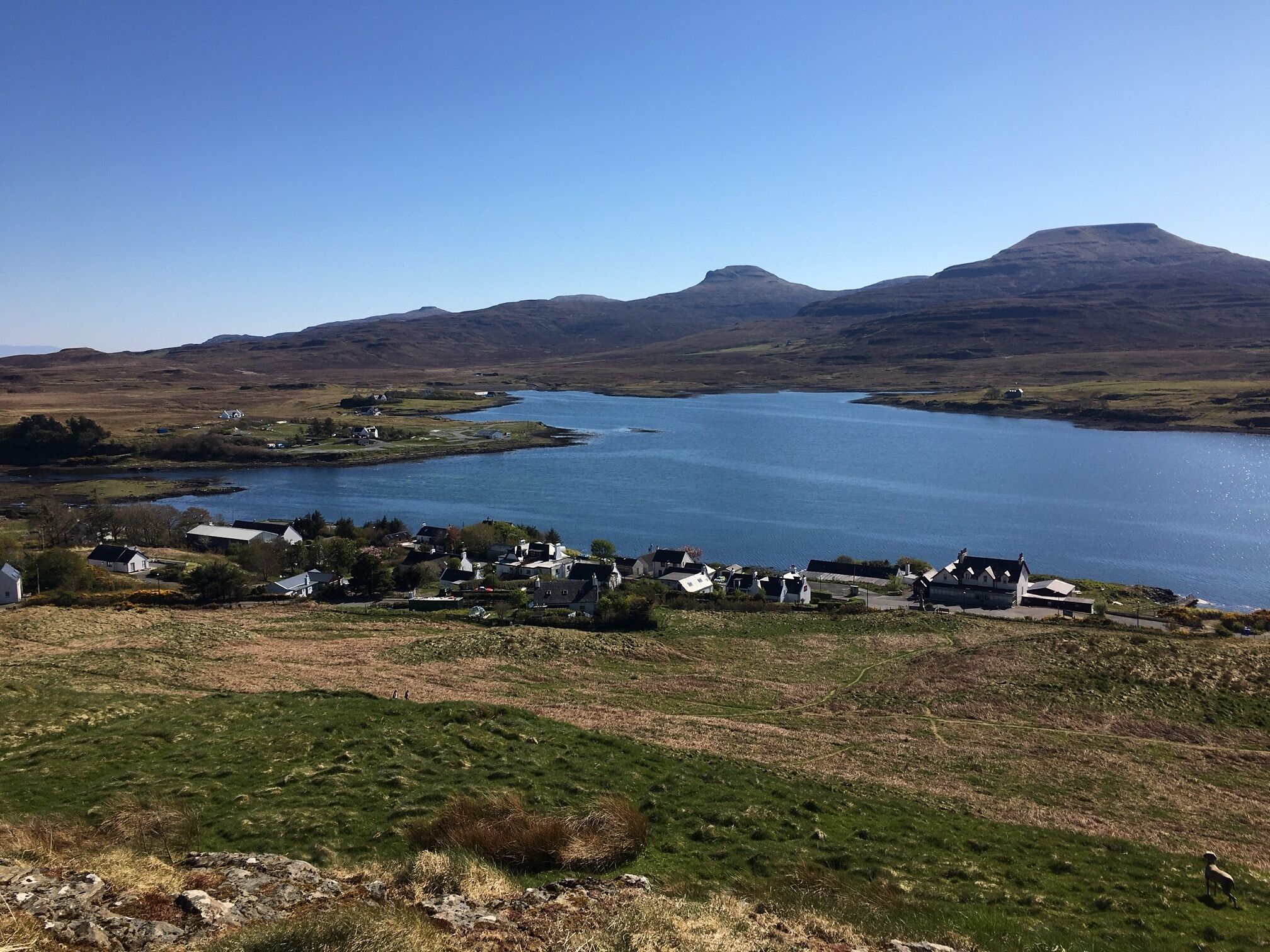 A coastal village with houses near a lake, surrounded by hills.