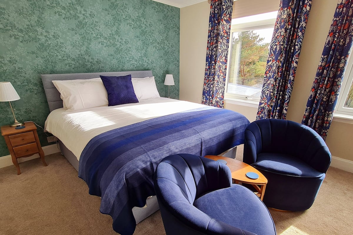 A bedroom with a bed, two chairs, a nightstand, and a window with curtains.