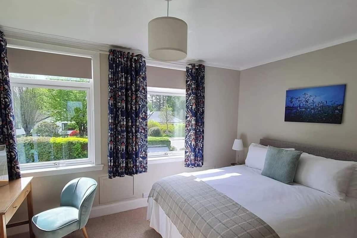 A bedroom with a bed, a desk, and a chair. There are two windows with curtains, a lamp, and a painting on the wall.