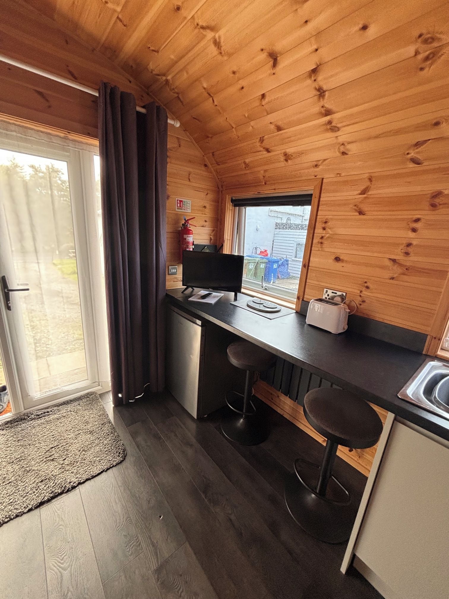 A compact kitchen with a wooden ceiling, a window with a view, a fire extinguisher, and a toaster.