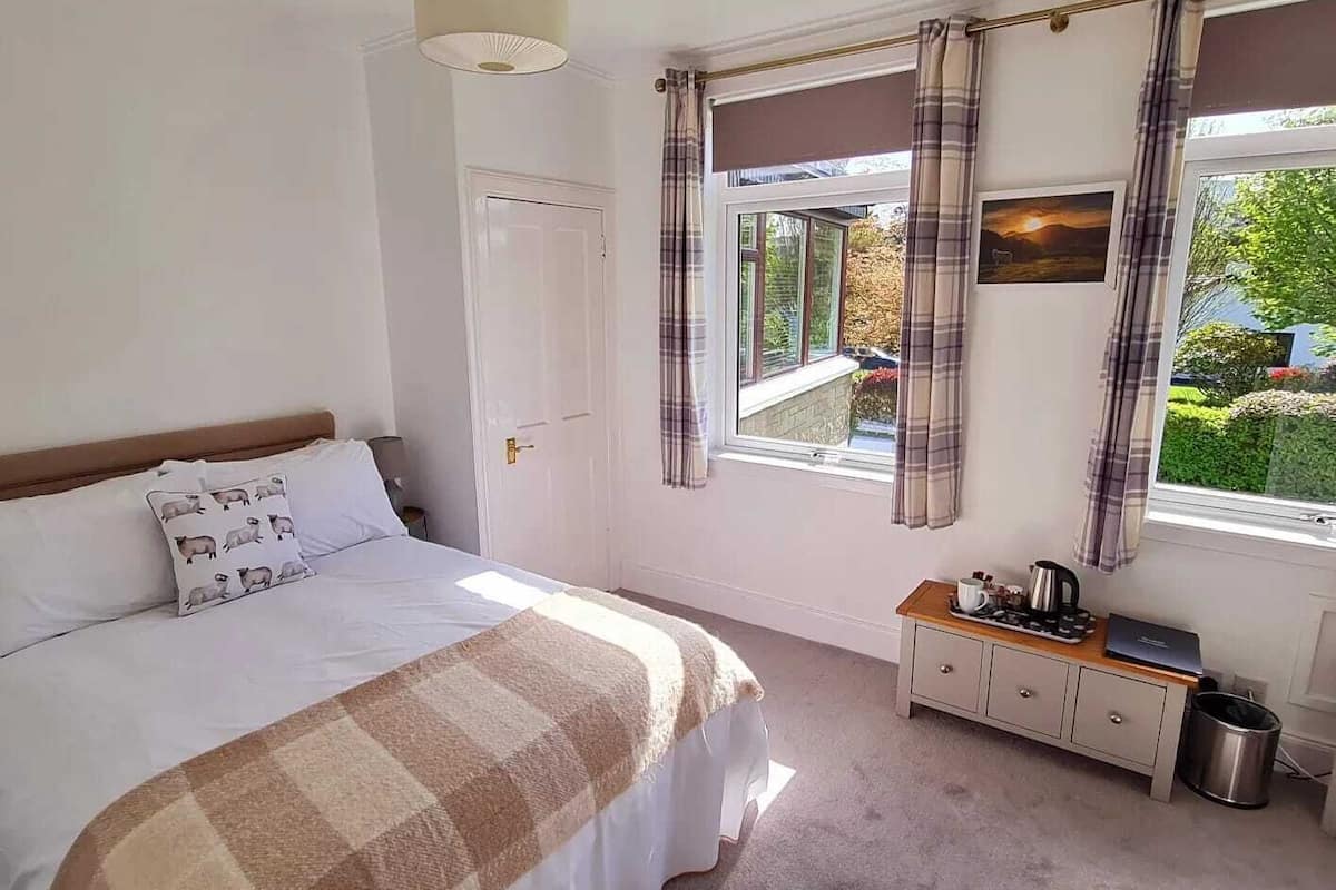 A bedroom with a bed, a bedside table with a kettle, a window with curtains, and a door.