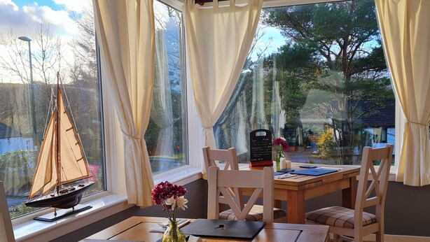A dining area with a wooden table set for two, a small sailboat model on the windowsill, and a menu board visible through the window. - Atholl House Skye (Dunvegan)