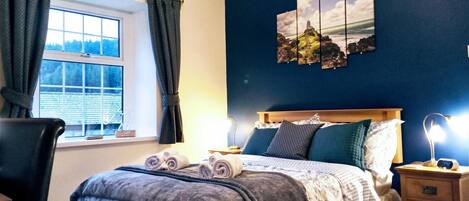 A bedroom with a bed, a nightstand, a chair, a window with curtains, and a wall with a landscape painting.