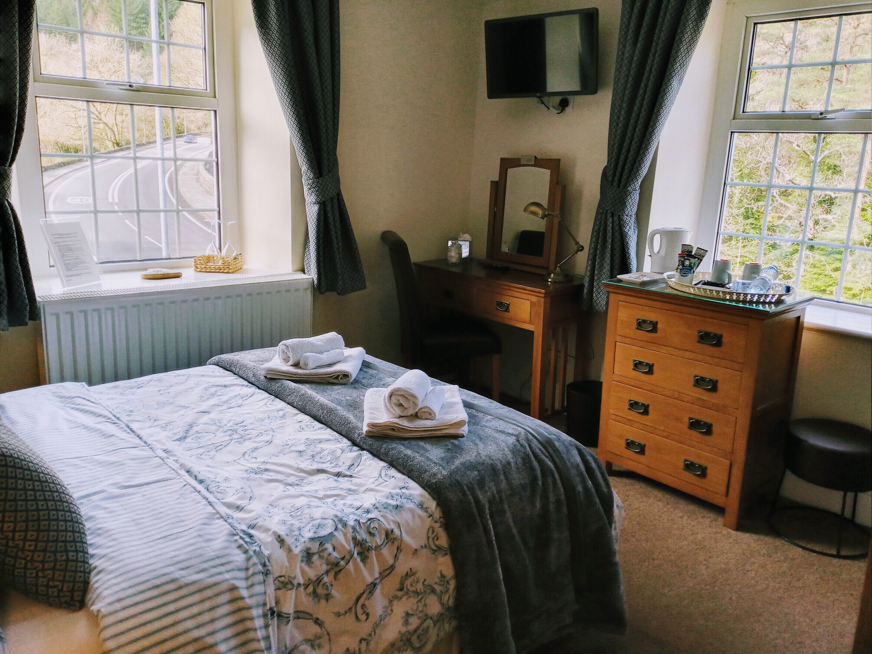 A neatly made bed with a patterned bedspread, a dresser with drawers, a mirror, and a television on the wall.
