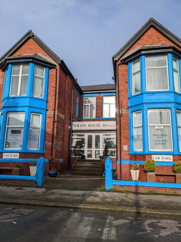 A Willin House Hotel with a traditional brick facade and blue trim. It features a central entrance with steps, a sign indicating a free car park, and a sign for the hotel on the ground floor. - Willin House Hotel (Blackpool)