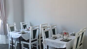 Un espace repas avec des tables et des chaises blanches, une nappe blanche et une fenêtre avec des rideaux.