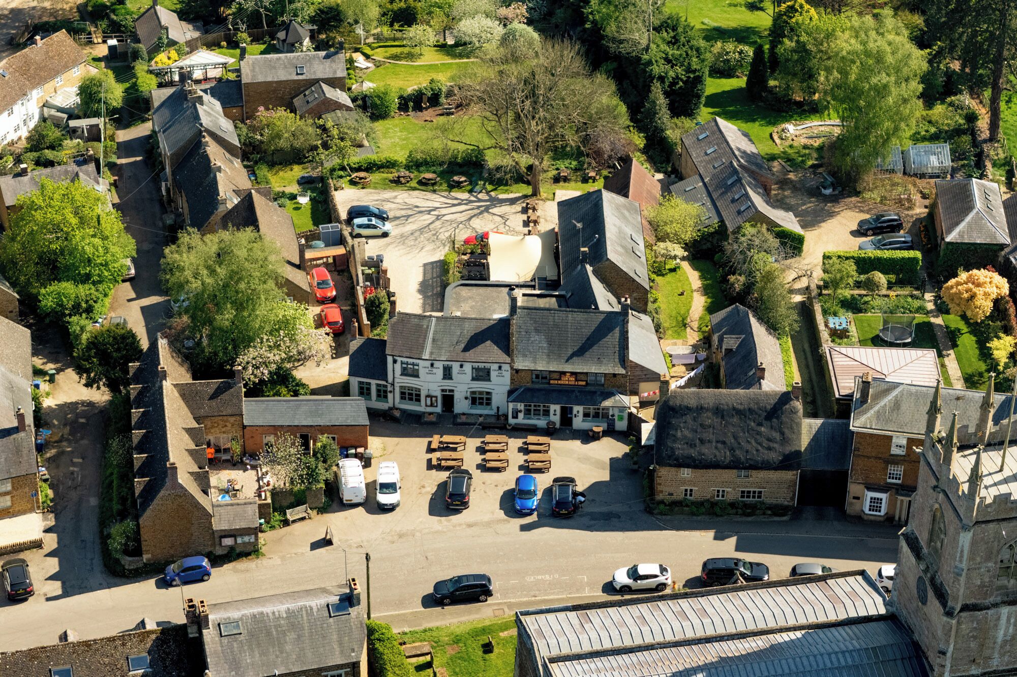 An aerial view of a village with a central pub, surrounded by residential buildings and a parking area.