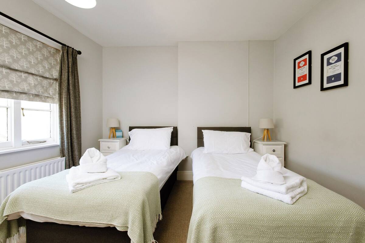 Quarto de hotel com duas camas de solteiro, roupa de cama branca, radiador, janela com cortinas e duas quadros na parede.