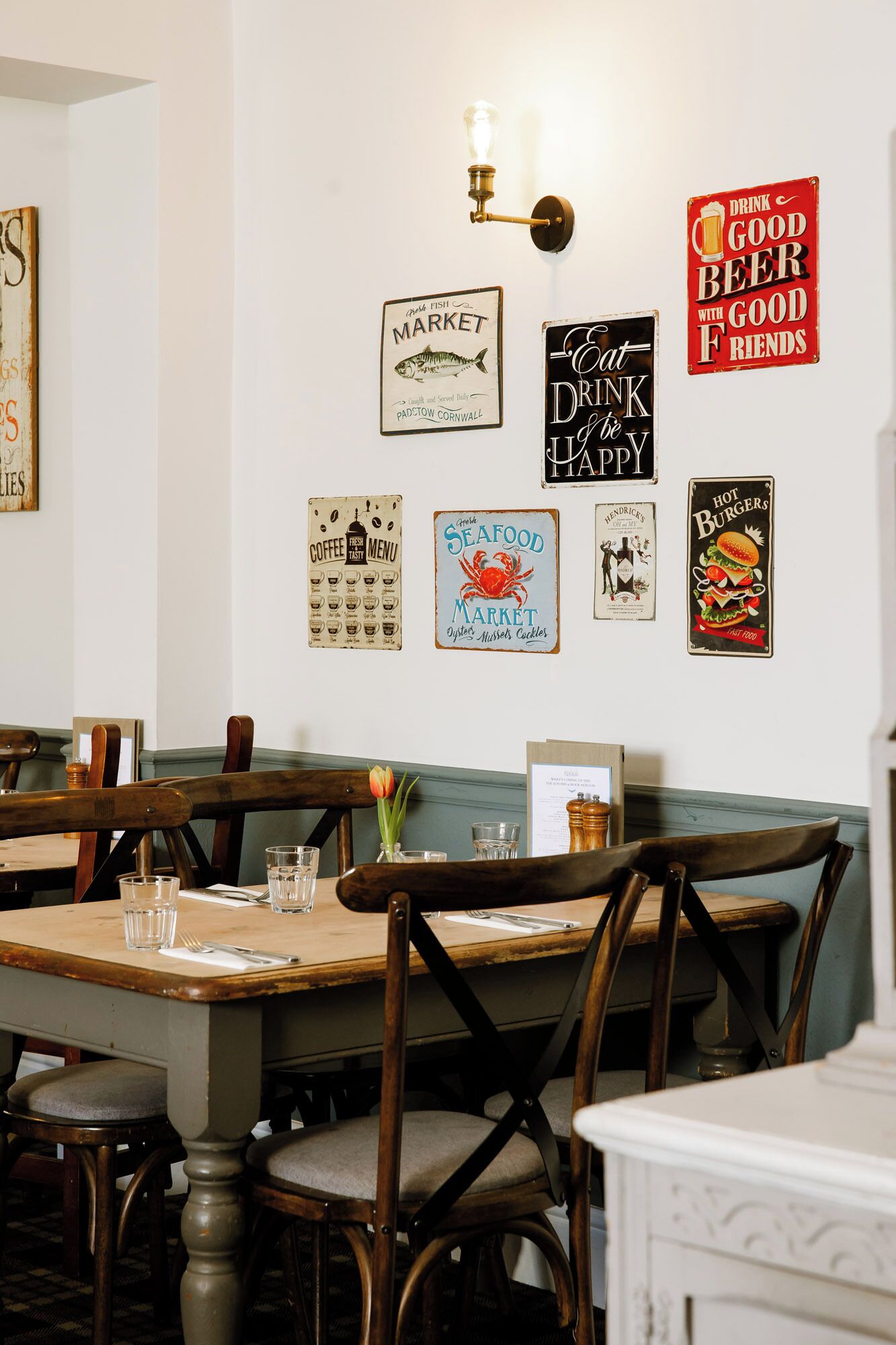 Un interior de restaurante con mesas y sillas de madera, una pared decorada con carteles de estilo vintage, y una lámpara que emite luz cálida.