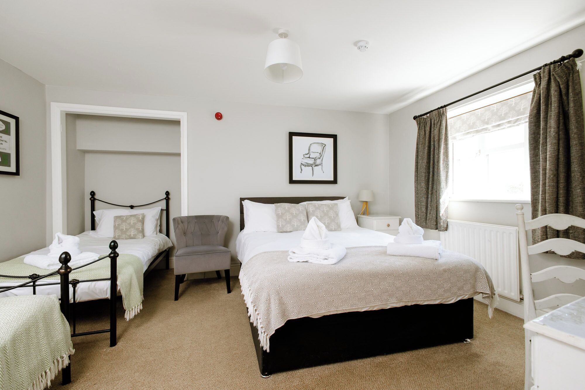 A hotel room with a large bed, two smaller beds, a chair, a desk, and a window with curtains.