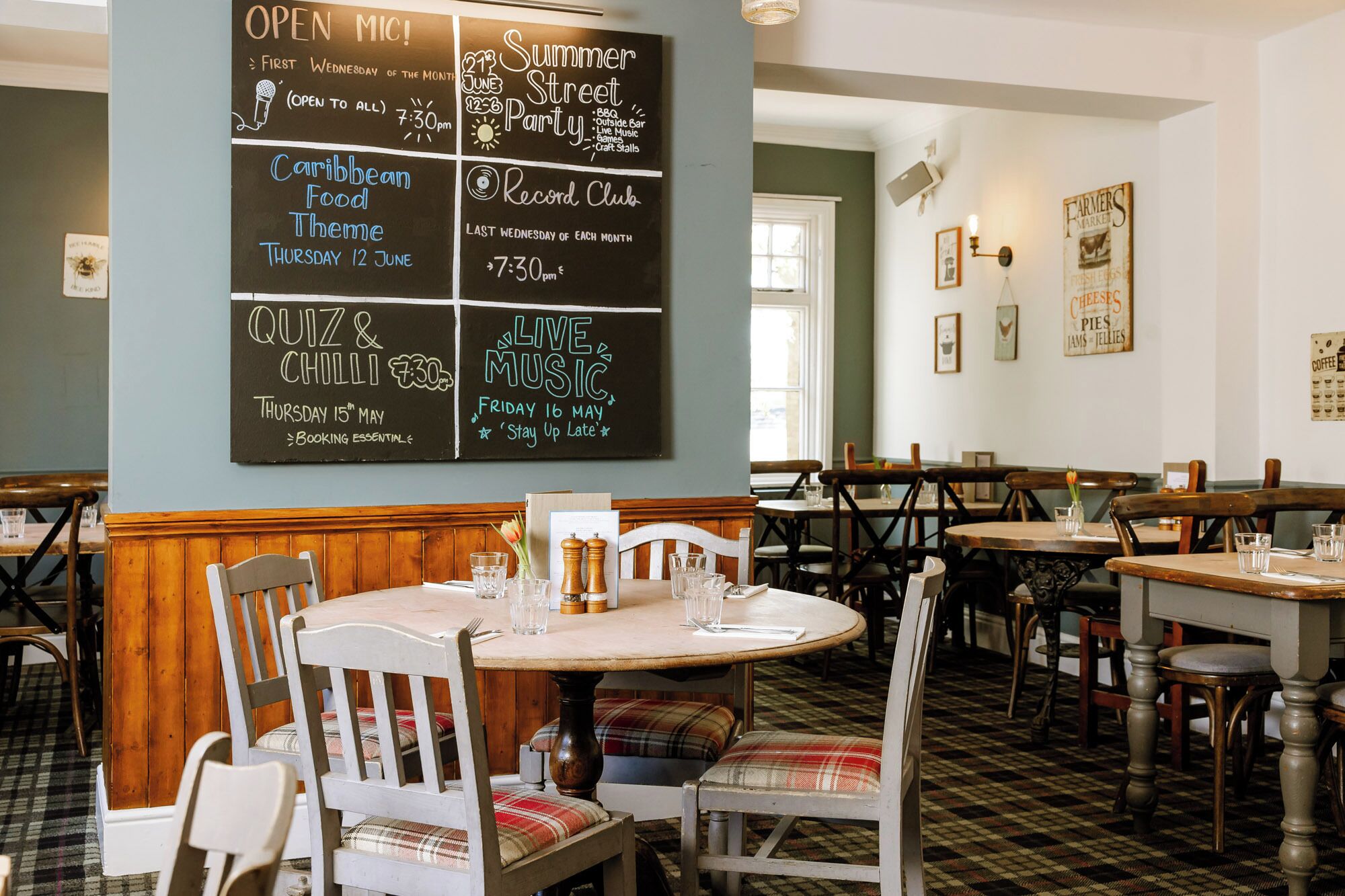 Interior de restaurante con un menú en pizarra que anuncia eventos como 'Open Mic' y 'Noche de comida caribeña'. Las mesas están dispuestas con sillas, y la decoración incluye elementos de madera y suelo estampado.