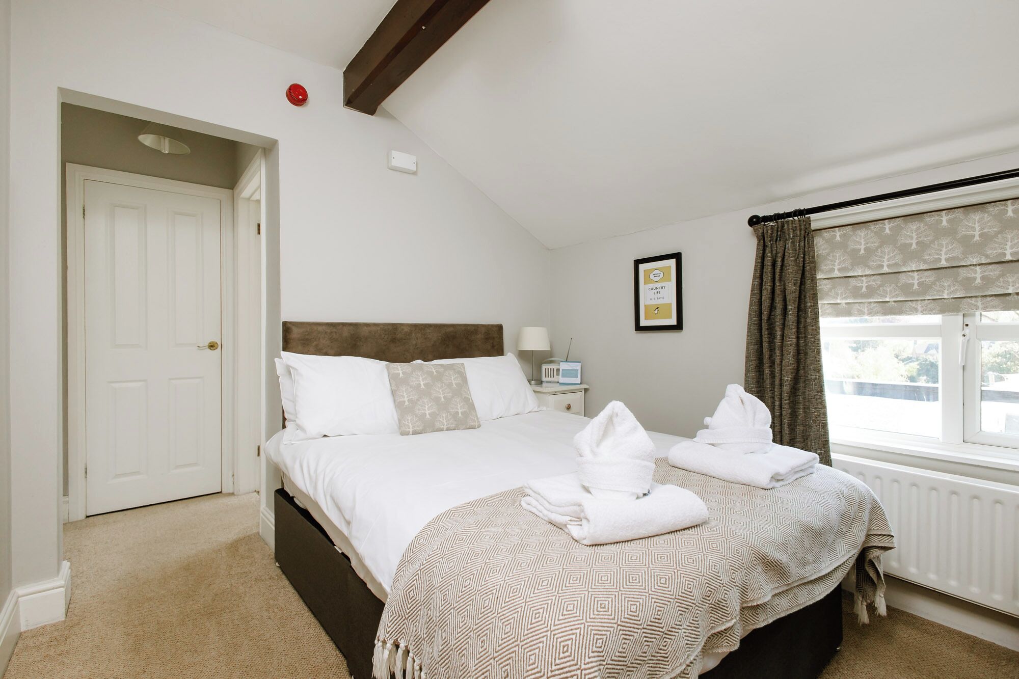 A bedroom with a large bed, a bedside table with a lamp, a framed picture on the wall, and a window with curtains.