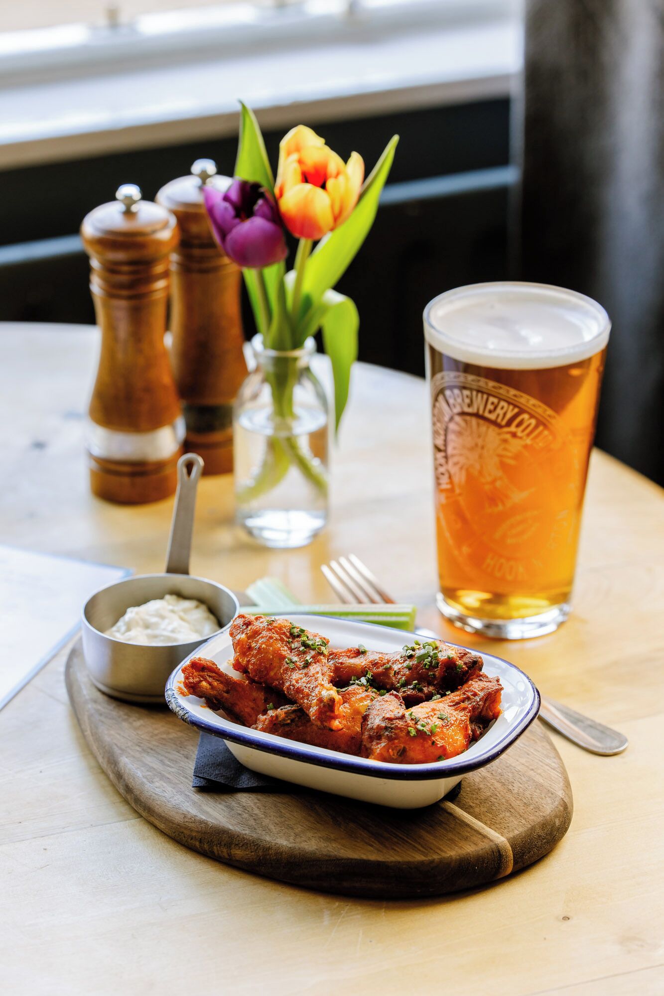 A meal with a side of sauce, a glass of beer, and a vase with flowers on a table.