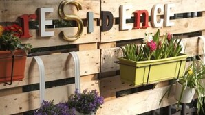 Pared de madera con la palabra "RESIDENCE" en letras grandes, plantas en macetas y una tubería blanca.