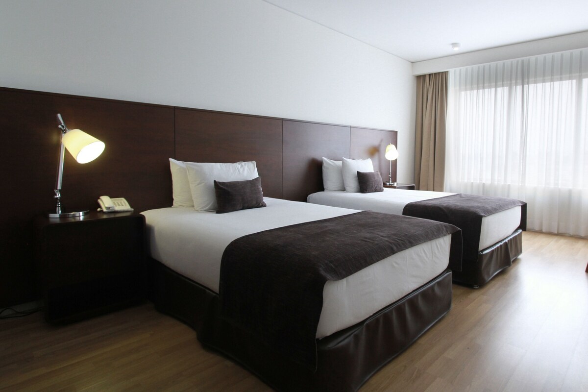 Una habitación de hotel con dos camas, un escritorio, una silla y un ventanal con cortinas.