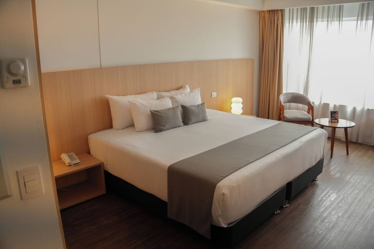 Ein Hotelzimmer mit einem großen Bett, einem Nachttisch mit Telefon, einem Sessel, einem kleinen Tisch und einem Fenster mit Vorhängen.