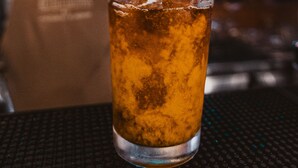 Een glas amberkleurig vocht met condens, een persoon giet in van een klein flesje, en een wazige achtergrond met planken en flessen.