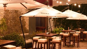 Een buitenterras met meerdere houten tafels en stoelen, waarvan sommige onder witte parasols met 'STELLA ARTOIS'-branding.