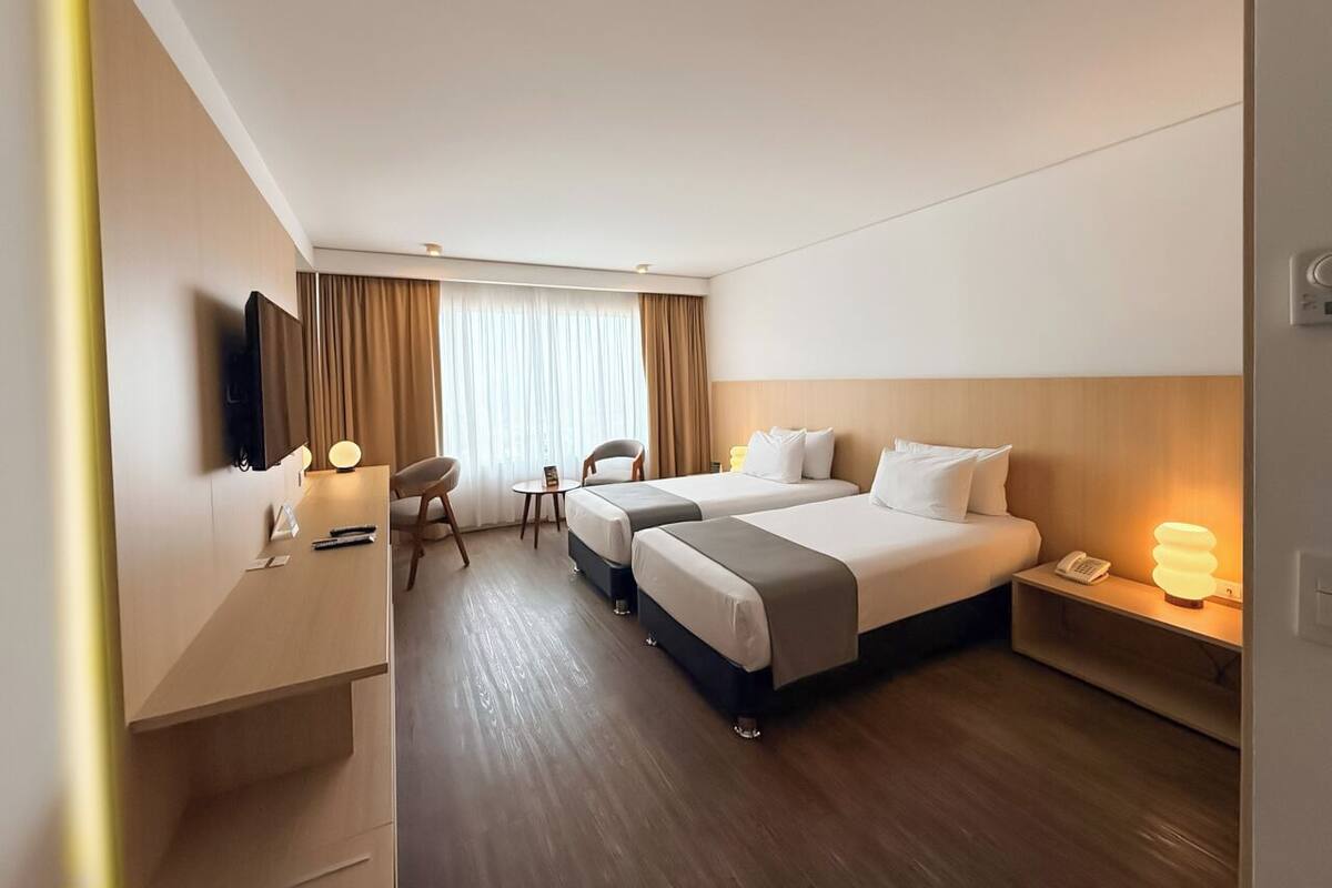Ein Hotelzimmer mit zwei Betten, einem Schreibtisch, einem Sessel, einem Fernseher und einem Nachttisch mit einer Lampe.