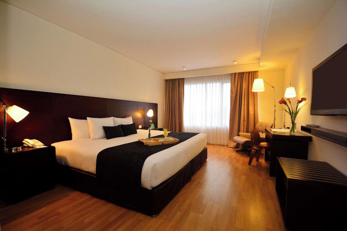 Ein Hotelzimmer mit einem großen Bett, Nachttischlampen, einem Schreibtisch mit Stuhl, einem Fernseher und einem Fenster mit Vorhängen.
