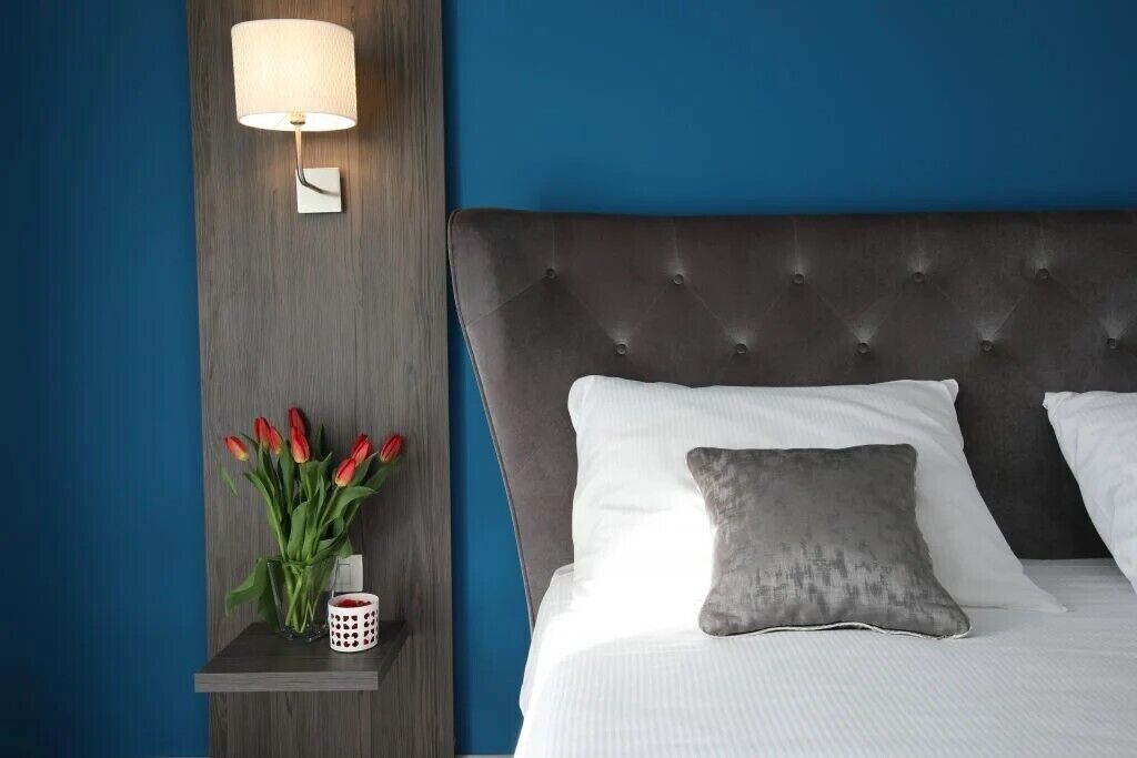 Un dormitorio con cabecera capitonada, ropa de cama blanca, un almohadón gris, una mesita de noche con una lámpara y un florero con tulipanes rojos.