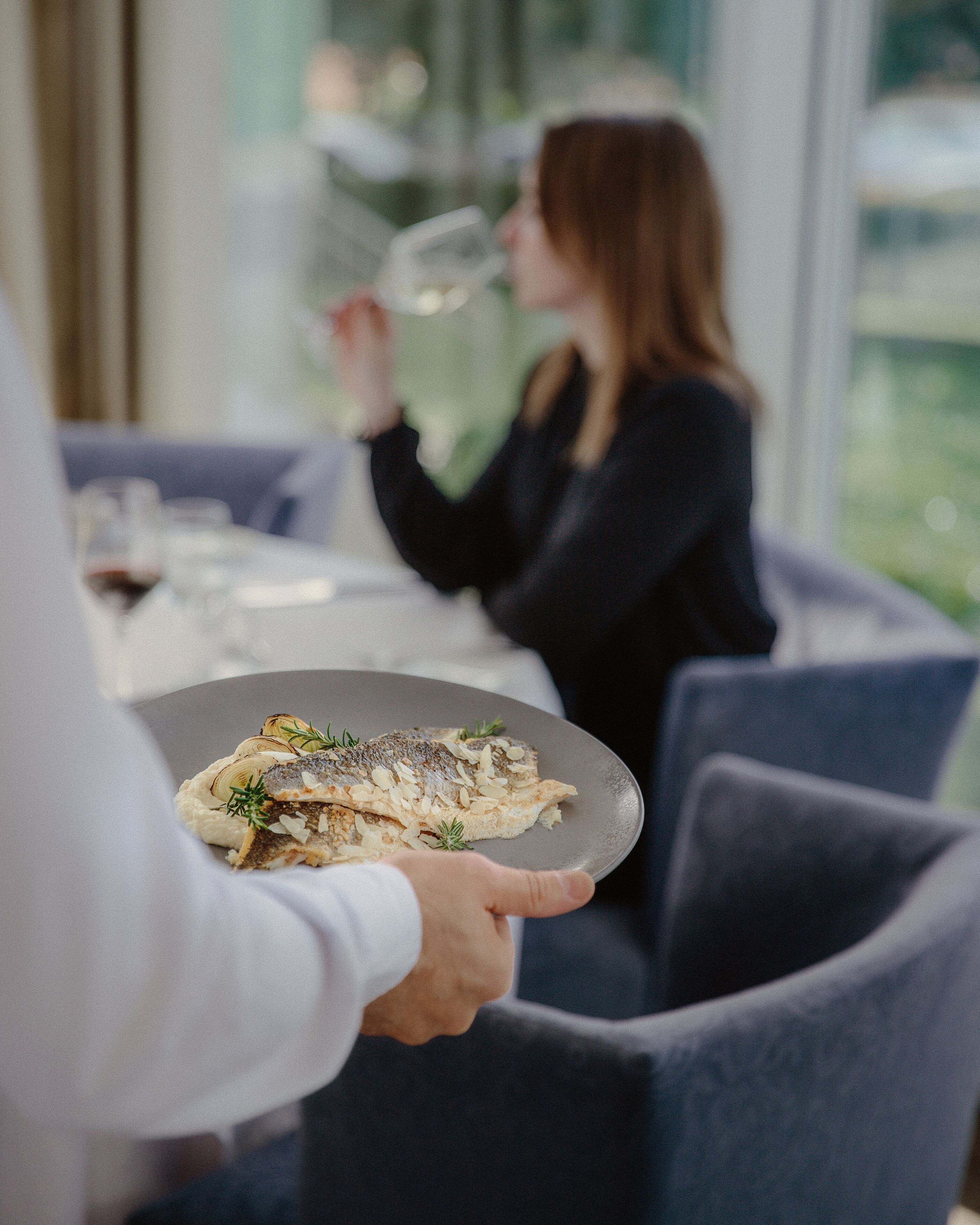 Eine Person serviert ein Fischgericht auf einem Teller an eine sitzende Person an einem Tisch, wo ein Glas Wein steht.