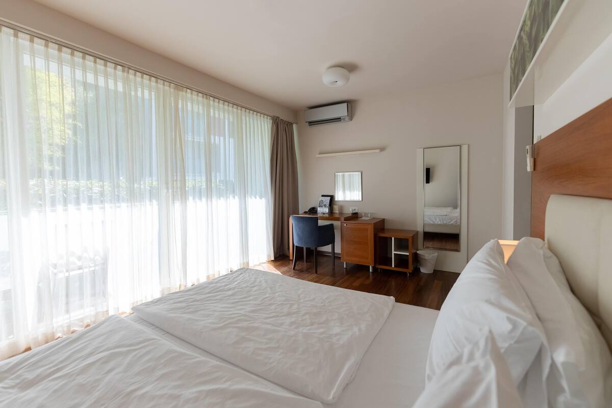 Habitación de hotel con una cama grande, un escritorio, una silla, una mesita y una ventana con cortinas translúcidas.