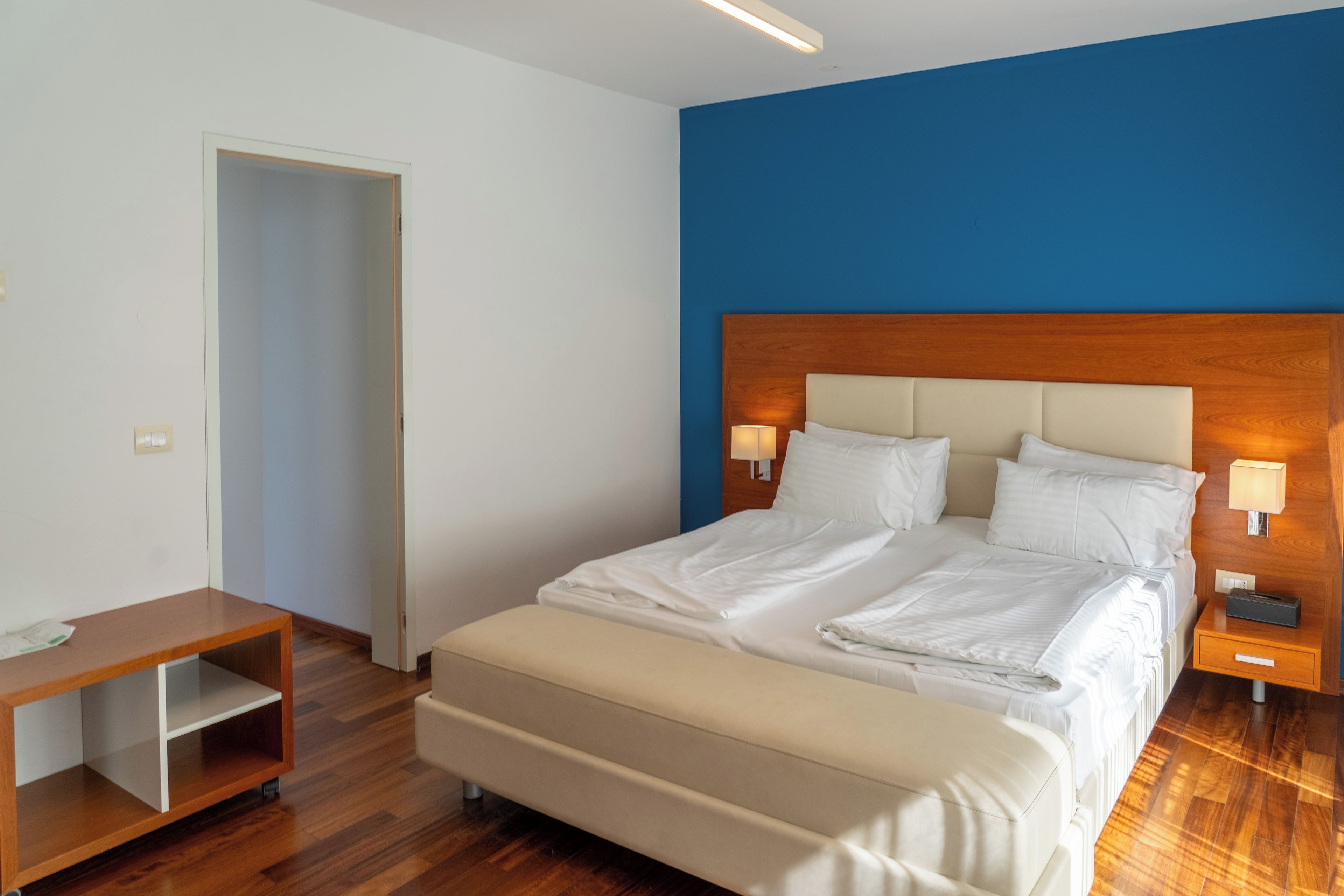 A hotel room with a bed, bedside lamps, a wooden nightstand, a small table, and a door.