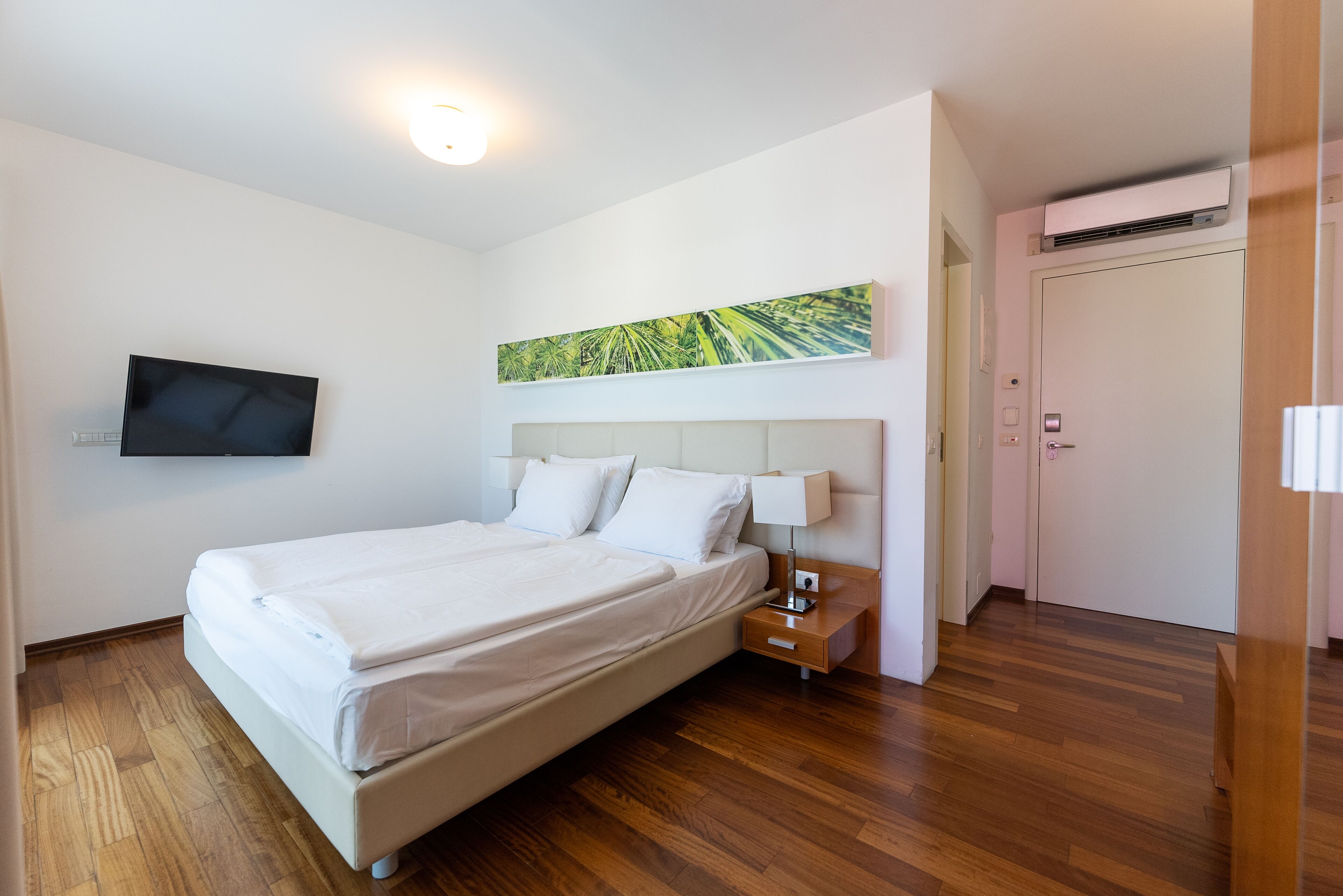 Ein ordentlich bezogenes Bett mit weißen Leinentüchern, ein Nachttisch mit Lampe, ein flachbildfernseher an der Wand und ein Gemälde über dem Bett.