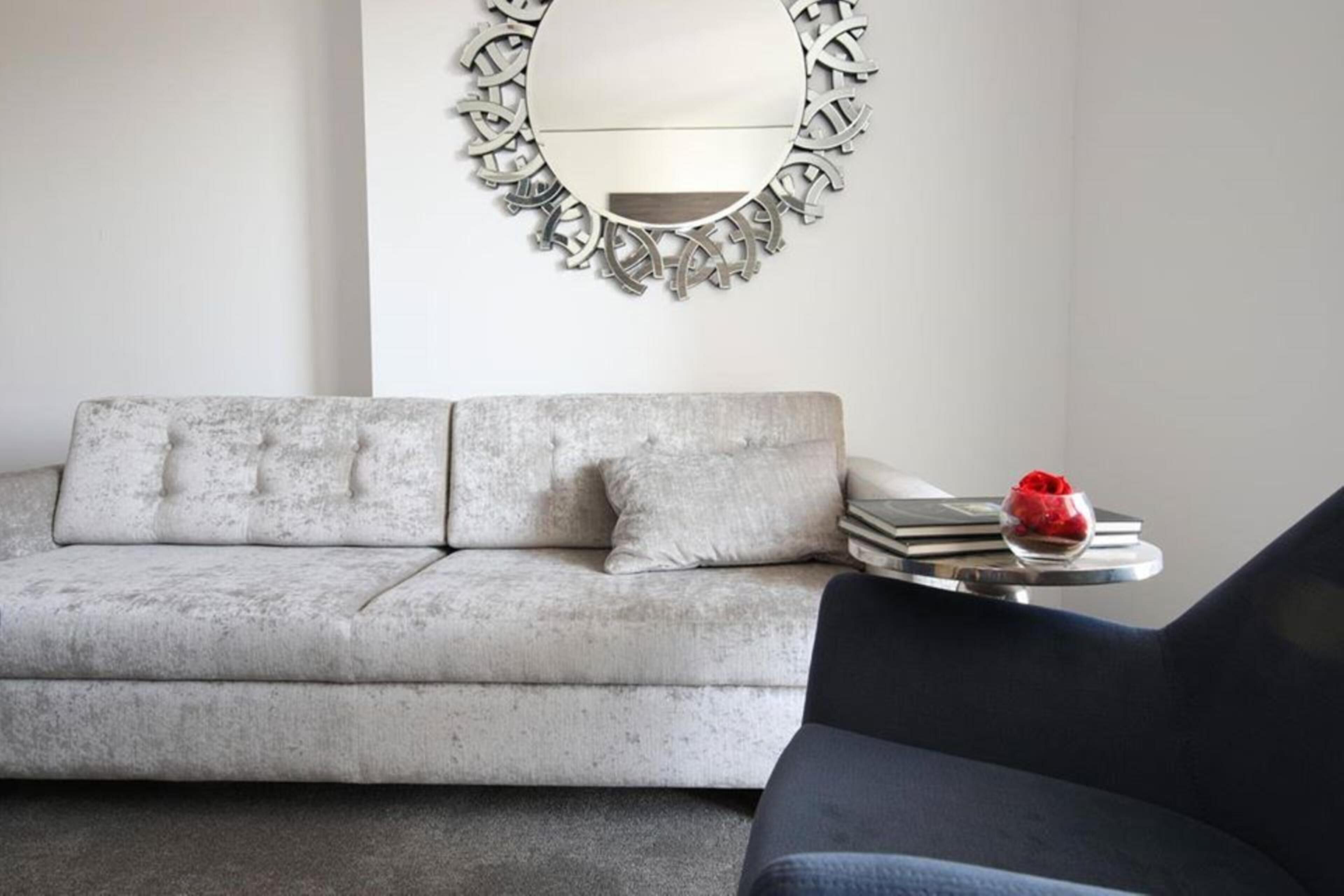 Ein Wohnzimmer mit grauem Sofa, einem runden Spiegel mit Metallrahmen und einem kleinen Tisch mit Büchern und einer roten Rose.