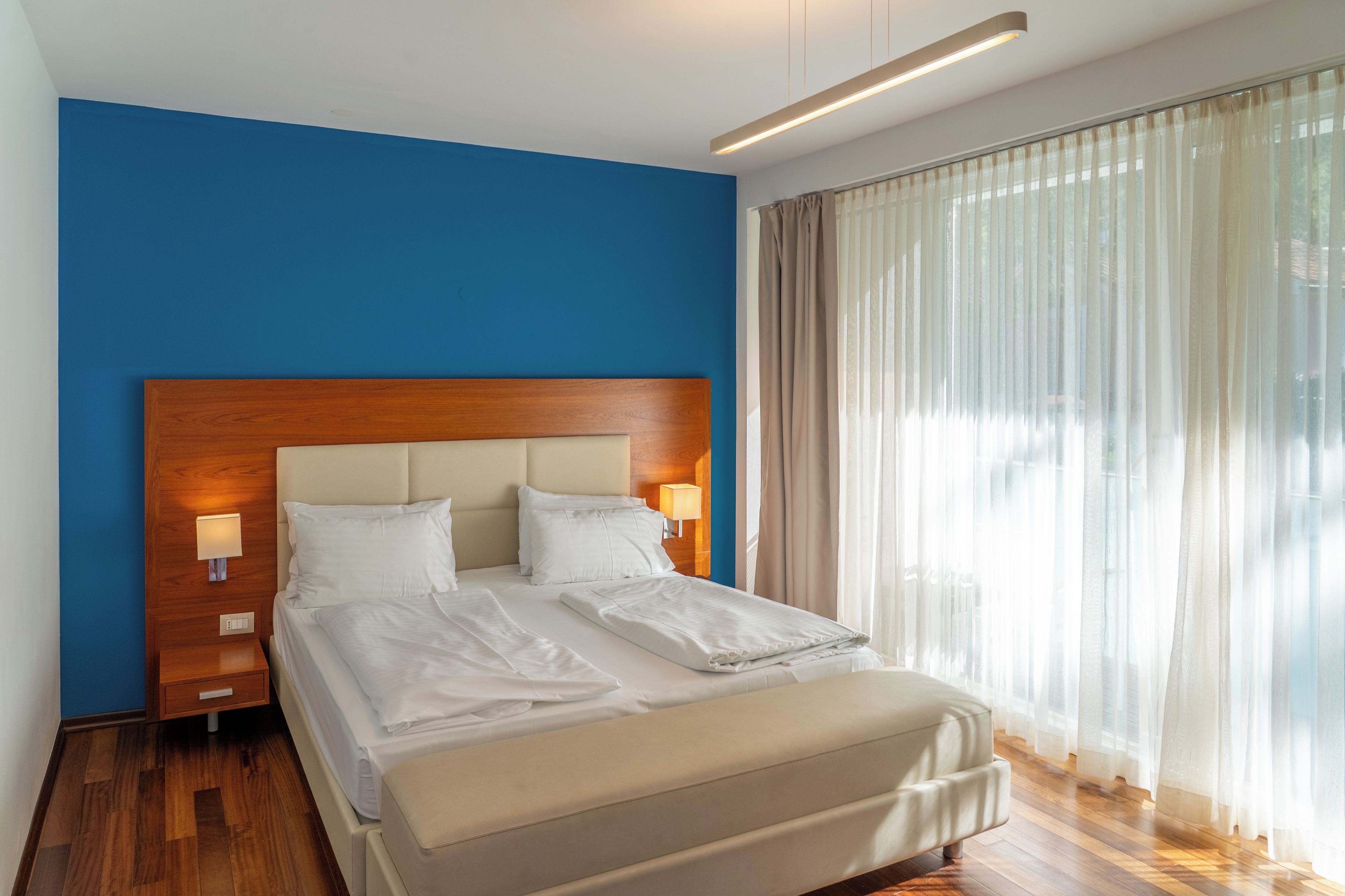 A hotel room with a large bed, wooden headboard, two bedside tables with lamps, and sheer curtains on the window.