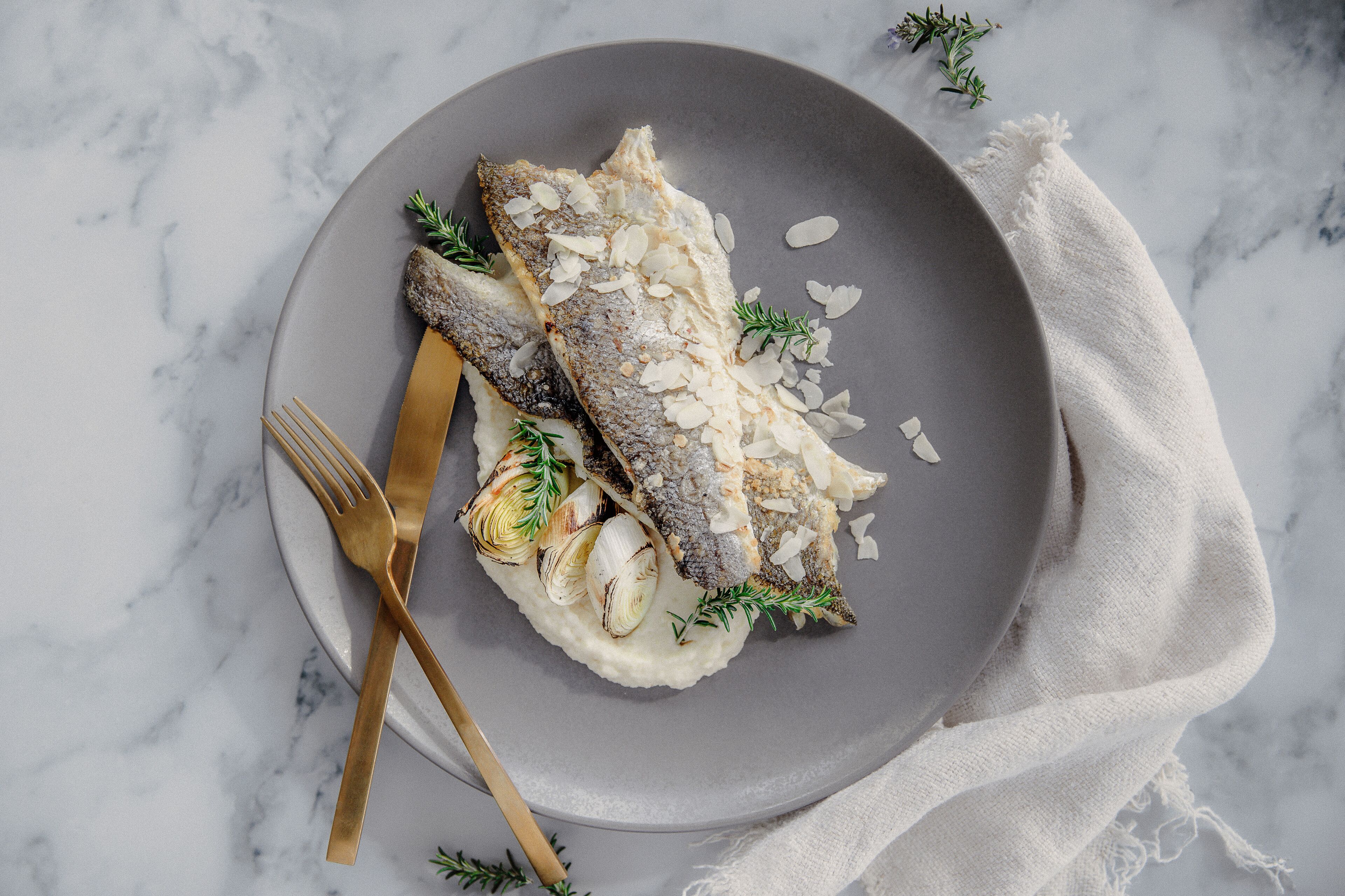 A plate with a fish, garnished with herbs and almonds, on a marble surface.