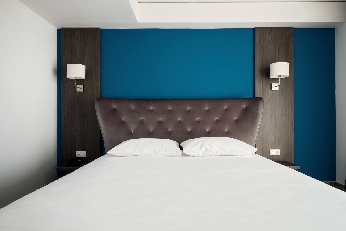 Una habitación de hotel moderna con cabecera capitonada, ropa de cama blanca y dos lámparas fijadas a la pared.