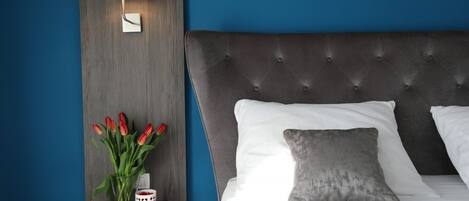 Un dormitorio con cabecera capitonada, ropa de cama blanca, un almohadón gris, una mesita de noche con un jarrón de tulipanes rojos y una lámpara de pared.