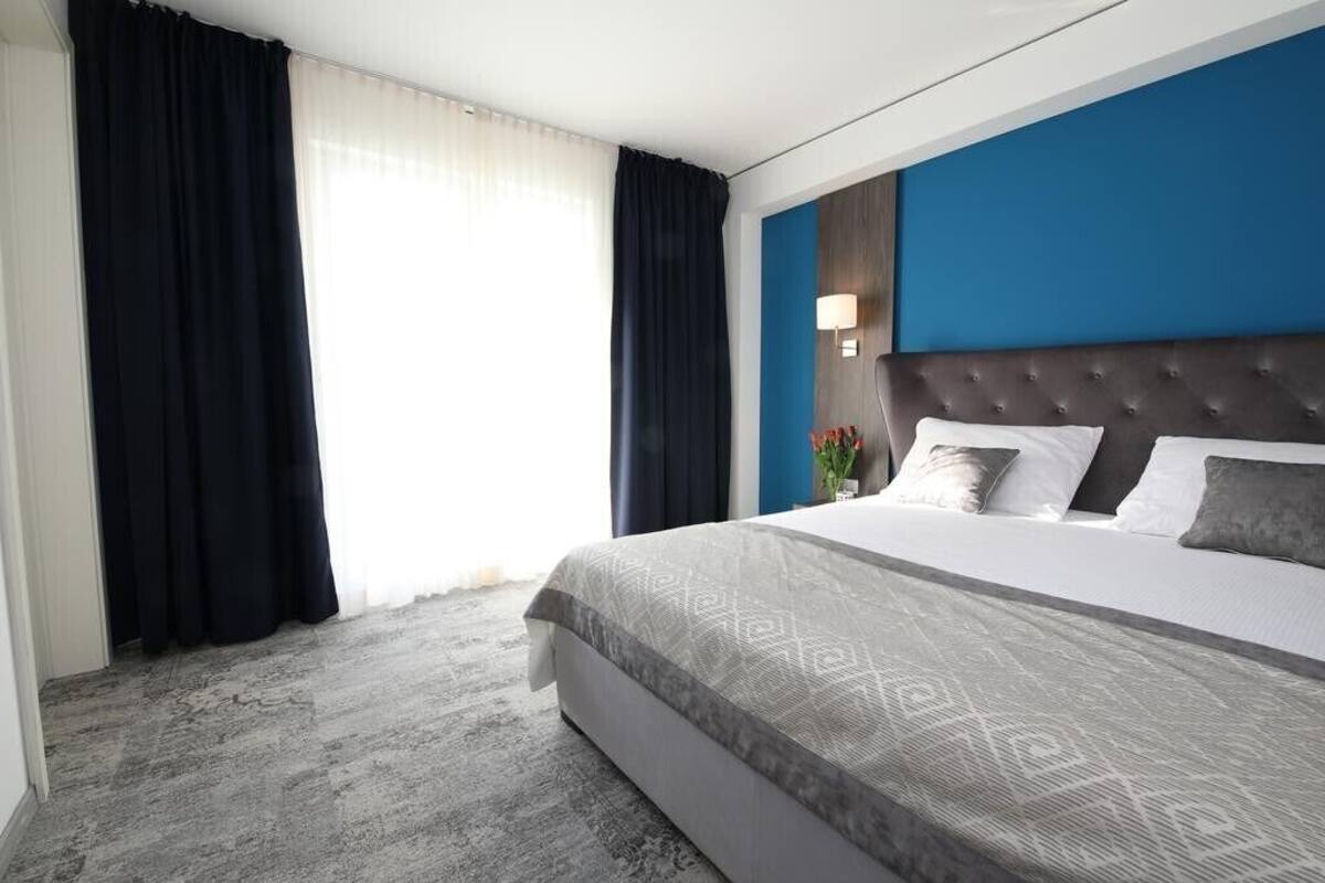 Una habitación de hotel moderna con una cama grande, cortinas oscuras, una pared azul como detalle y una colcha gris estampada.