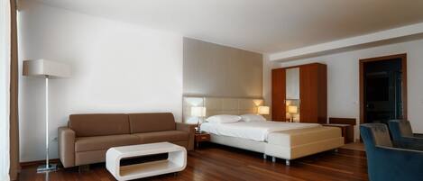 A modern hotel room with a wooden floor, a sofa, a bed, a desk, and a chair.