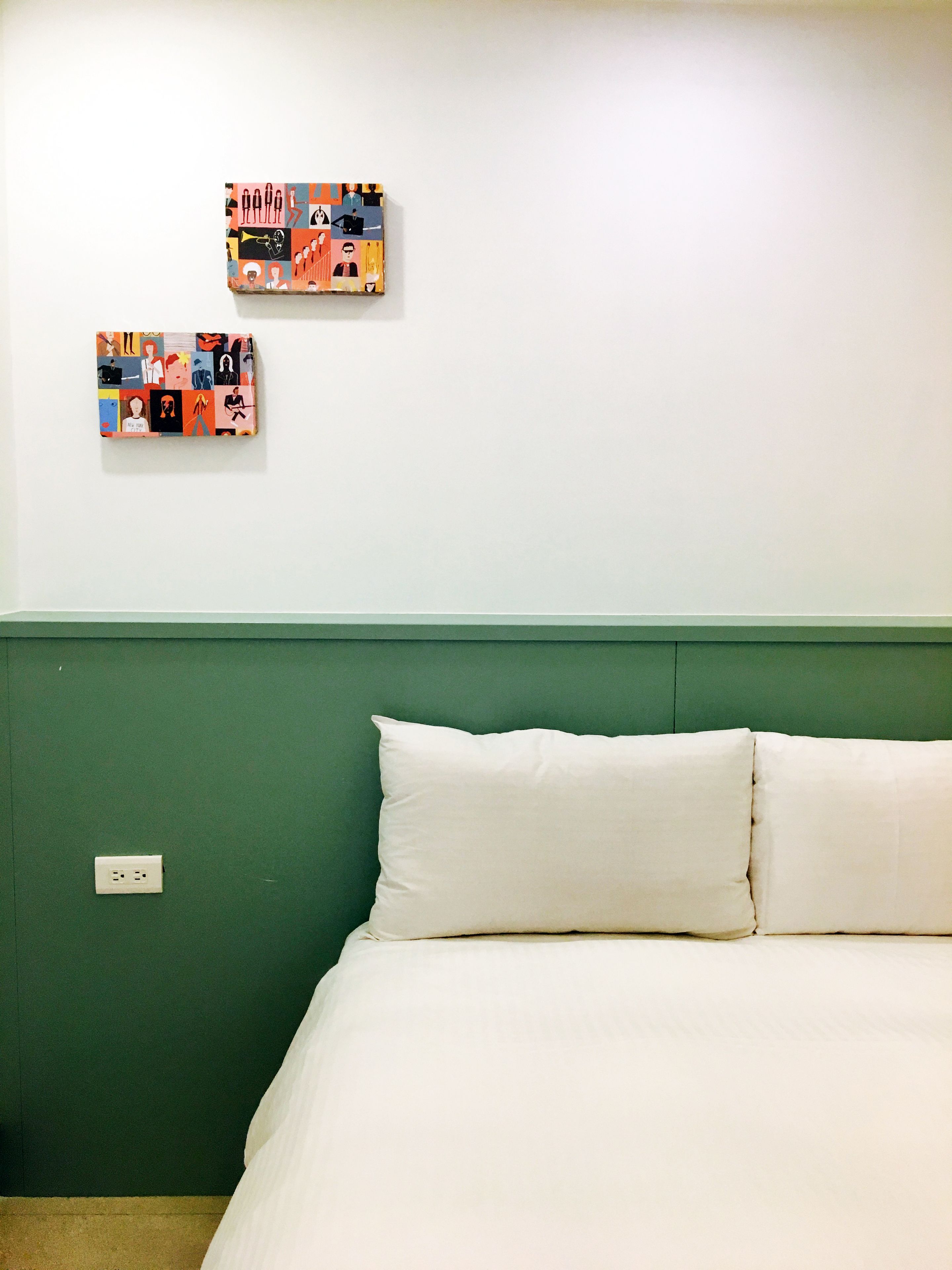 A room with a green wall, a white bed with two pillows, and two colorful artworks on the wall.