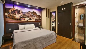 Una habitació d'hotel amb un llit gran, un quadre d'un poble europeu a la paret, un capçal de llit de fusta, i un bany amb plat de dutxa de vidre.